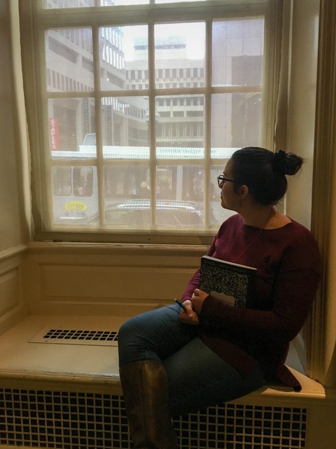 A young woman looks out a window with a composition notebook in hand.